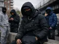 Młodzi mężczyźni w kapturach i maskach, w stylu drill, siedzą na murku. Jeden z nich ma na sobie czarną kurtkę puchową i torbę taktyczną.