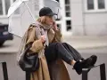 Kobieta w beżowym płaszczu, czapce i czarnych jeansach prezentuje modne emu stylizacje, trzymając przezroczysty parasol.