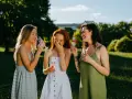 Trzy kobiety śmieją się na trawie z kieliszkami wina. Idealne inspiracje, w co się ubrać na wesele po 40.