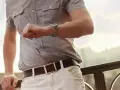 Mężczyzna w szarej koszuli i białych spodniach sprawdza zegarek. Czarny pasek z prostą klamrą idealnie podkreśla jego styl. Jak dobrać pasek?
