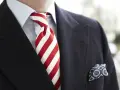 Elegancki mężczyzna w granatowym garniturze, błękitnej koszuli i z wyrazistym, czerwonym krawatem w paski. W kieszeni marynarki chusteczka w niebieskie wzory.