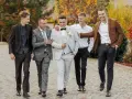 Pięciu mężczyzn w eleganckich, męskich stylizacjach na jesień, spaceruje po brukowanej ścieżce wśród jesiennych liści.