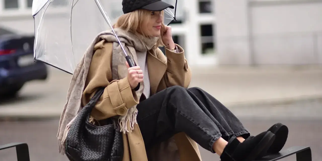 Kobieta w beżowym płaszczu, czapce i czarnych jeansach prezentuje modne emu stylizacje, trzymając przezroczysty parasol.