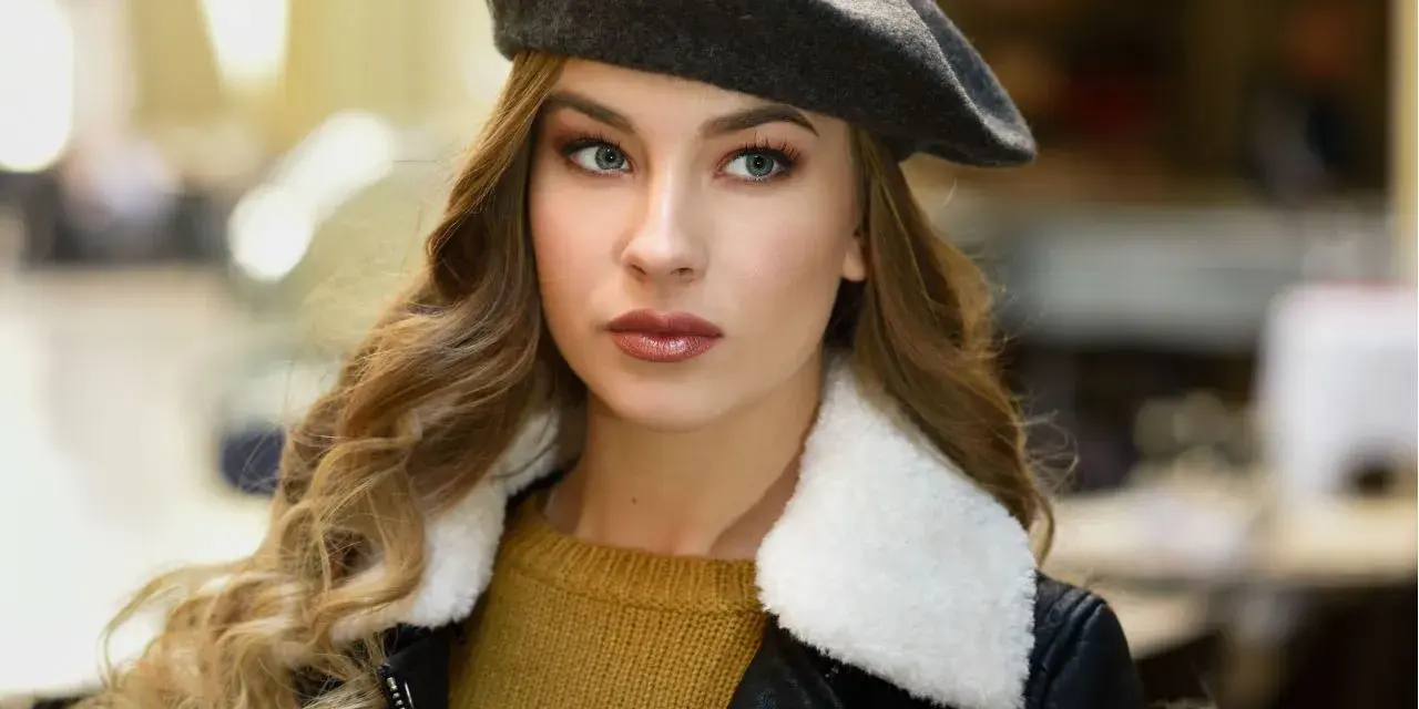 Elegancka kobieta w grafitowym beretcie, pokazująca, jak się nosi beret, z burzą blond loków i w stylowej kurtce.