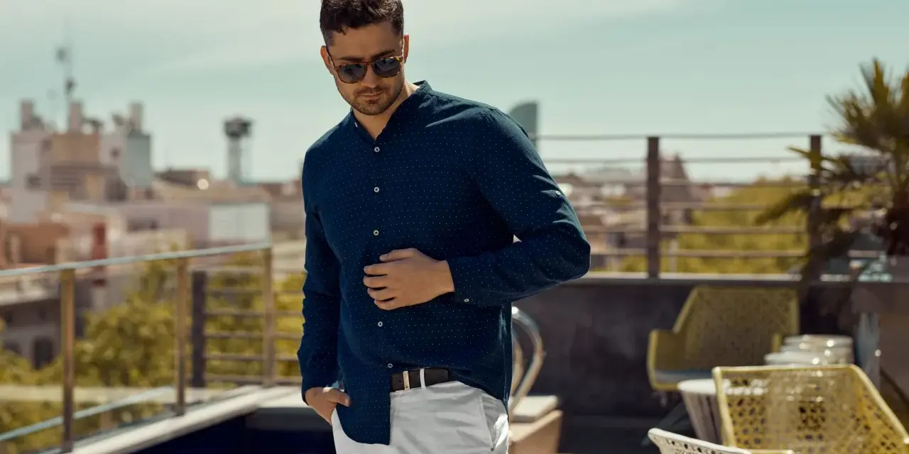 Mężczyzna w granatowej koszuli w kropki i białych spodniach, prezentujący luźny elegancki styl męski na tarasie z widokiem na miasto.
