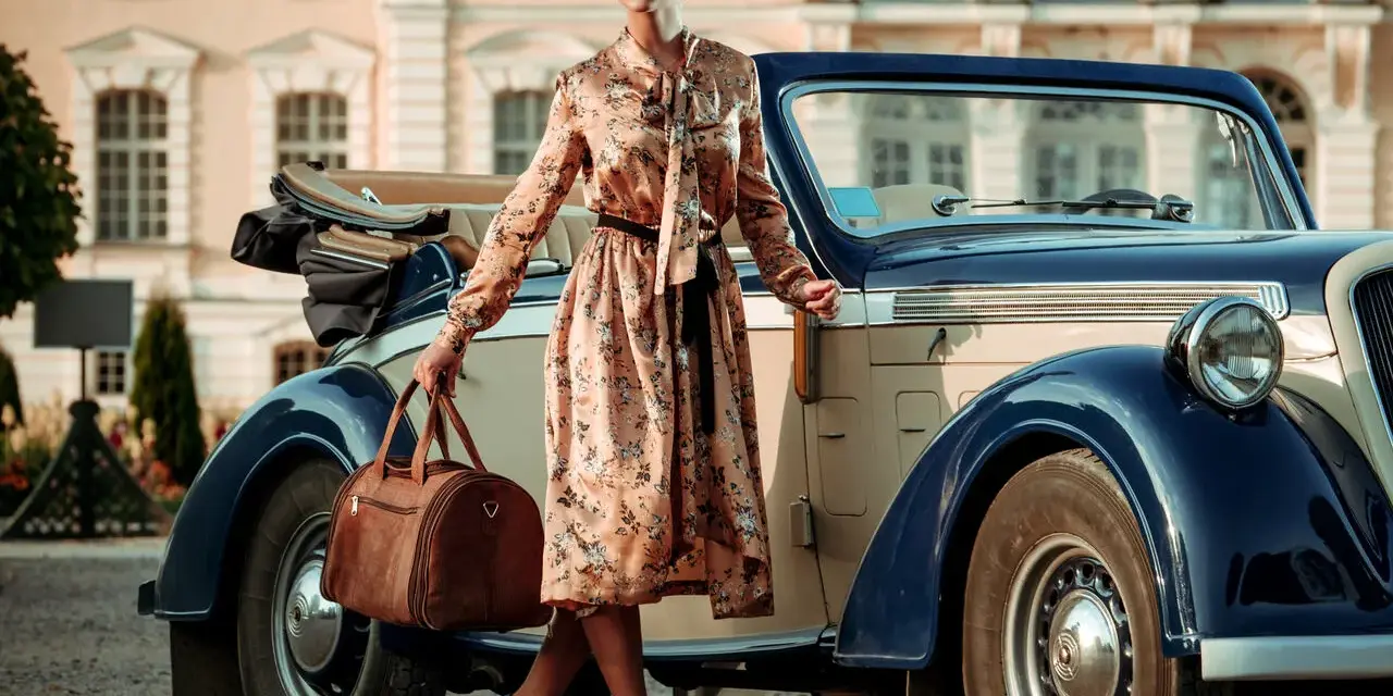 Elegancka kobieta w kwiecistej sukience i okularach przeciwsłonecznych, z brązową torbą podróżną, przy zabytkowym samochodzie. Styl vintage moda damska.