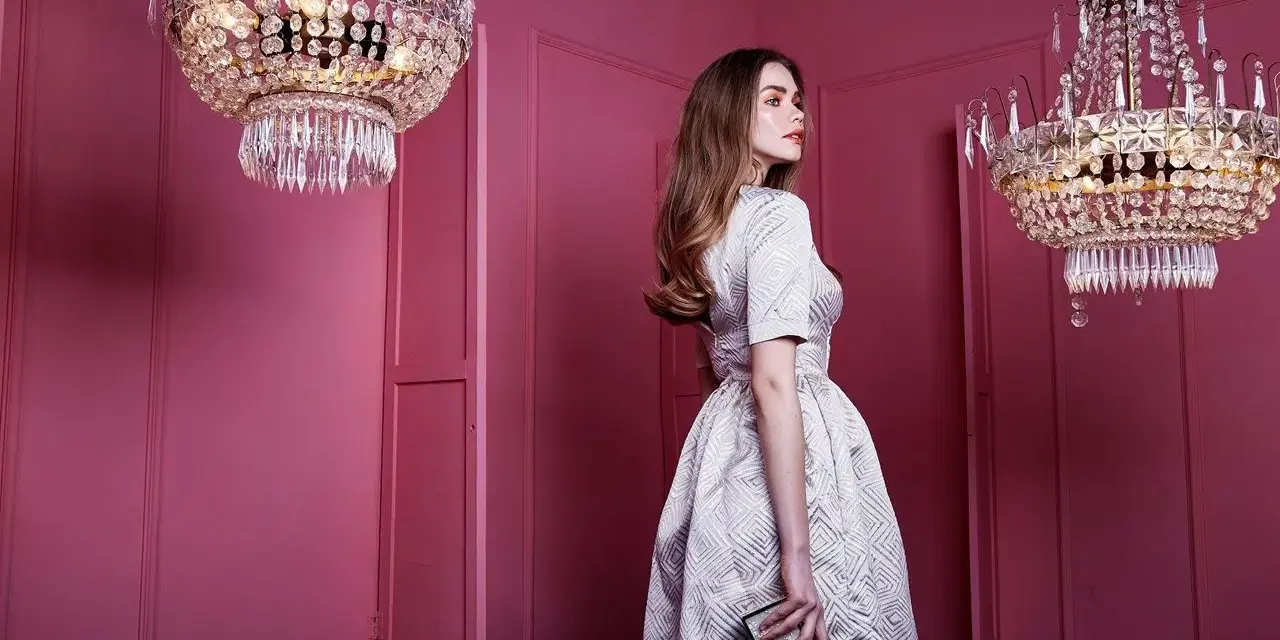 Elegancka kobieta w sukience, idealna na sukienki dla mamy pana młodego, pozuje przy kryształowych żyrandolach w różowym wnętrzu.