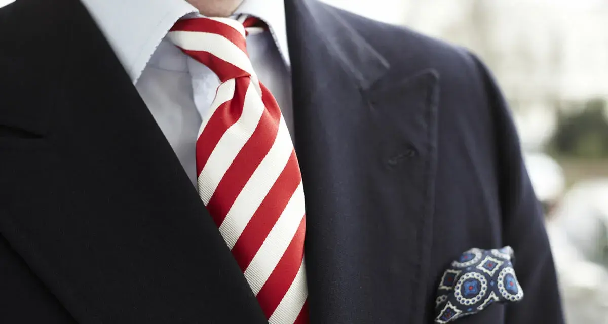 Elegancki mężczyzna w granatowym garniturze, błękitnej koszuli i z wyrazistym, czerwonym krawatem w paski. W kieszeni marynarki chusteczka w niebieskie wzory.