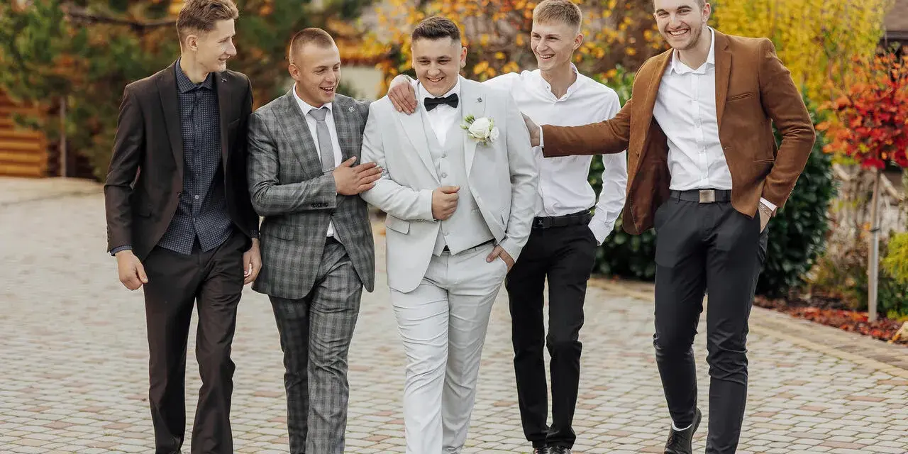 Pięciu mężczyzn w eleganckich, męskich stylizacjach na jesień, spaceruje po brukowanej ścieżce wśród jesiennych liści.