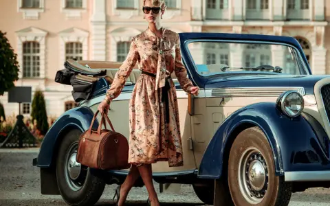 Elegancka kobieta w kwiecistej sukience i okularach przeciwsłonecznych, z brązową torbą podróżną, przy zabytkowym samochodzie. Styl vintage moda damska.