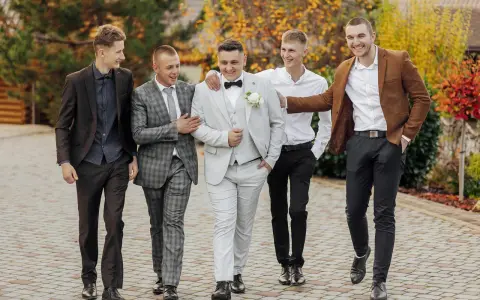 Pięciu mężczyzn w eleganckich, męskich stylizacjach na jesień, spaceruje po brukowanej ścieżce wśród jesiennych liści.