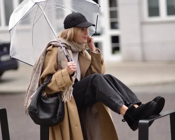 Kobieta w beżowym płaszczu, czapce i czarnych jeansach prezentuje modne emu stylizacje, trzymając przezroczysty parasol.