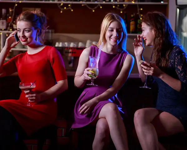 Trzy kobiety w strojach koktajlowych siedzą przy barze, pijąc drinki i rozmawiając.