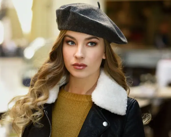Elegancka kobieta w grafitowym beretcie, pokazująca, jak się nosi beret, z burzą blond loków i w stylowej kurtce.