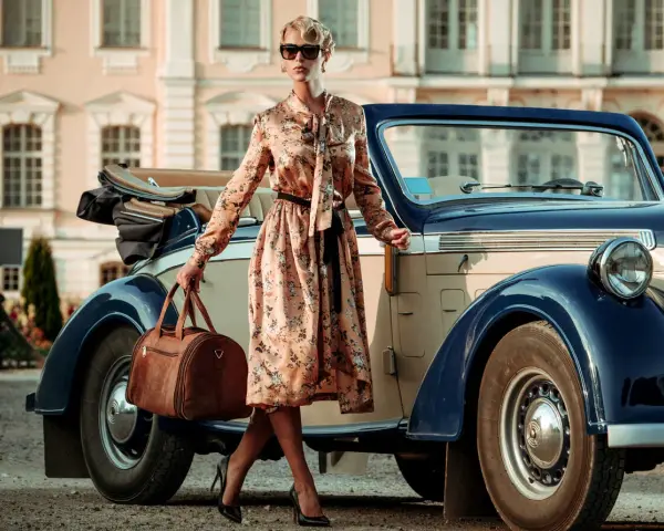 Elegancka kobieta w kwiecistej sukience i okularach przeciwsłonecznych, z brązową torbą podróżną, przy zabytkowym samochodzie. Styl vintage moda damska.