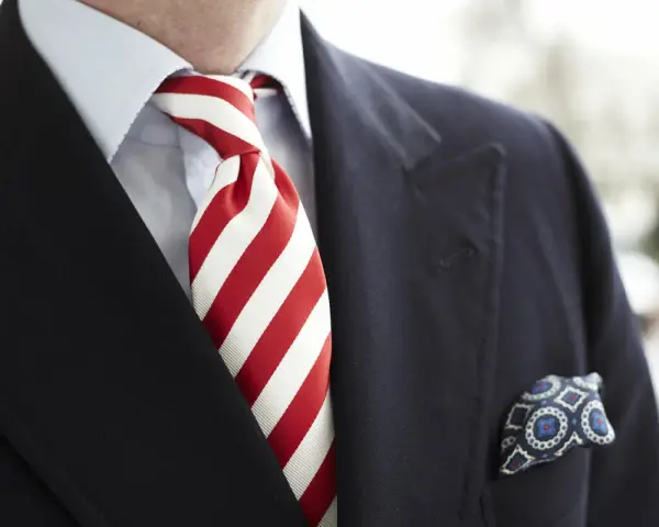 Elegancki mężczyzna w granatowym garniturze, błękitnej koszuli i z wyrazistym, czerwonym krawatem w paski. W kieszeni marynarki chusteczka w niebieskie wzory.