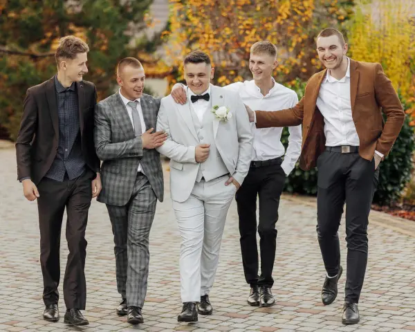 Pięciu mężczyzn w eleganckich, męskich stylizacjach na jesień, spaceruje po brukowanej ścieżce wśród jesiennych liści.