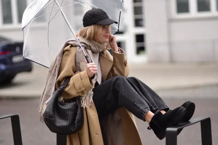 Kobieta w beżowym płaszczu, czapce i czarnych jeansach prezentuje modne emu stylizacje, trzymając przezroczysty parasol.