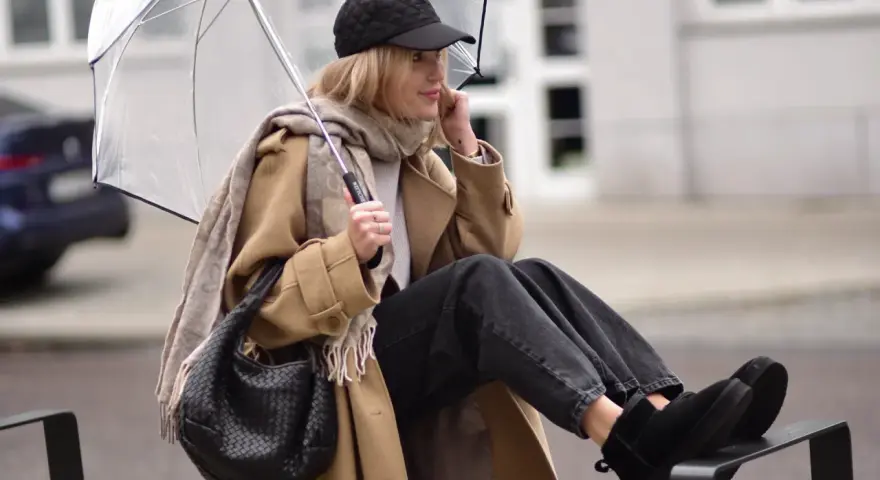 Kobieta w beżowym płaszczu, czapce i czarnych jeansach prezentuje modne emu stylizacje, trzymając przezroczysty parasol.