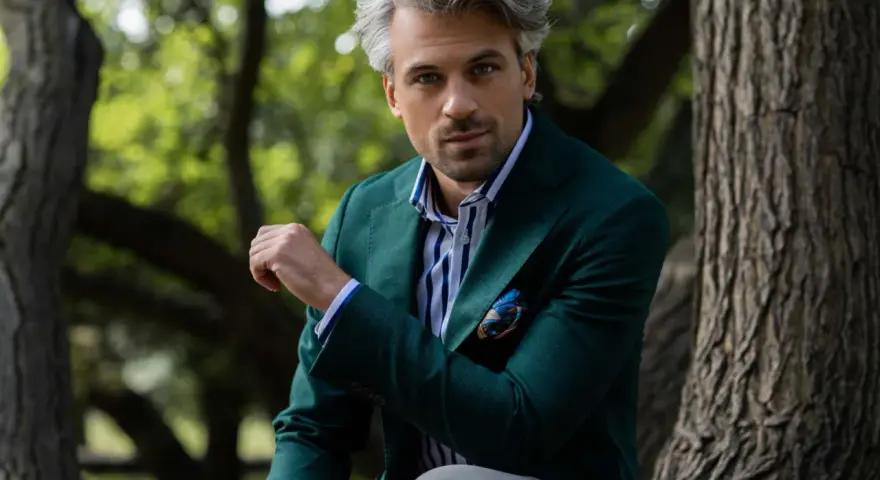 Elegancki mężczyzna w zielonej marynarce i koszulce polo z niebieskimi paskami, siedzący na tle drzew.
