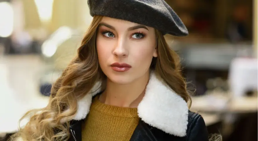 Elegancka kobieta w grafitowym beretcie, pokazująca, jak się nosi beret, z burzą blond loków i w stylowej kurtce.
