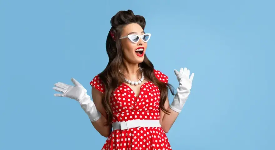 Urocza pin up girl w czerwonej sukience w grochy, białych rękawiczkach i okularach w kształcie kocich oczu, z uniesionymi rękami w geście zdziwienia.