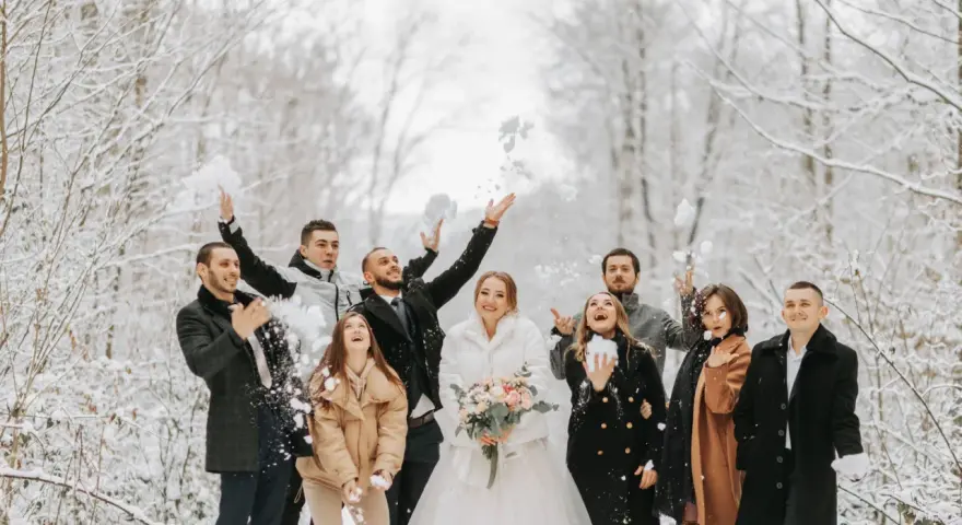 Radosna para młoda i goście rzucają śnieżkami na tle zimowego lasu. Idealne stylizacje na wesele zimą.