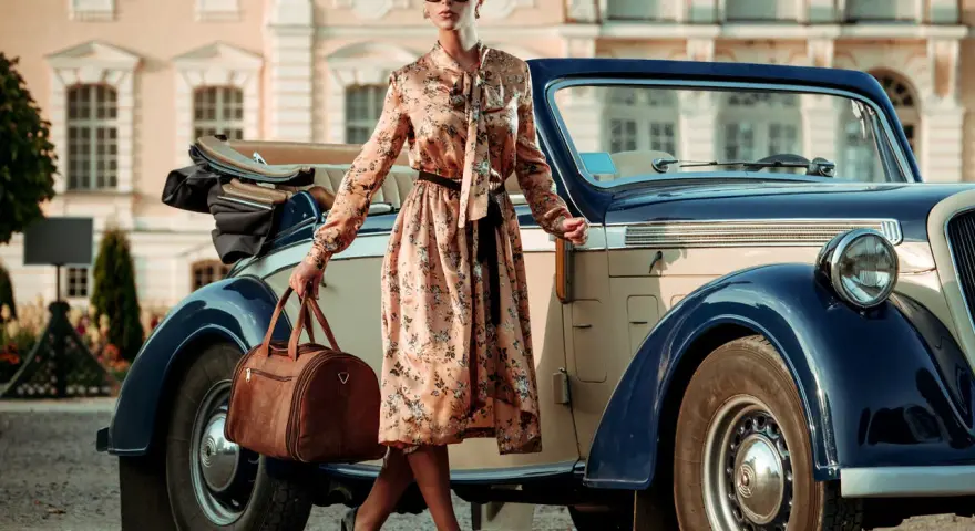 Elegancka kobieta w kwiecistej sukience i okularach przeciwsłonecznych, z brązową torbą podróżną, przy zabytkowym samochodzie. Styl vintage moda damska.