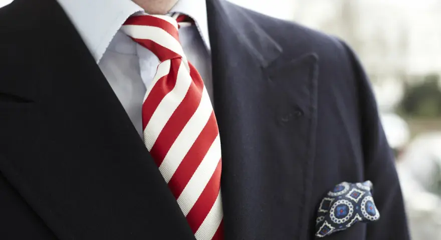 Elegancki mężczyzna w granatowym garniturze, błękitnej koszuli i z wyrazistym, czerwonym krawatem w paski. W kieszeni marynarki chusteczka w niebieskie wzory.