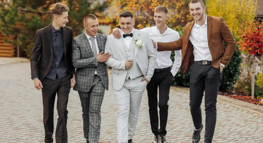Pięciu mężczyzn w eleganckich, męskich stylizacjach na jesień, spaceruje po brukowanej ścieżce wśród jesiennych liści.
