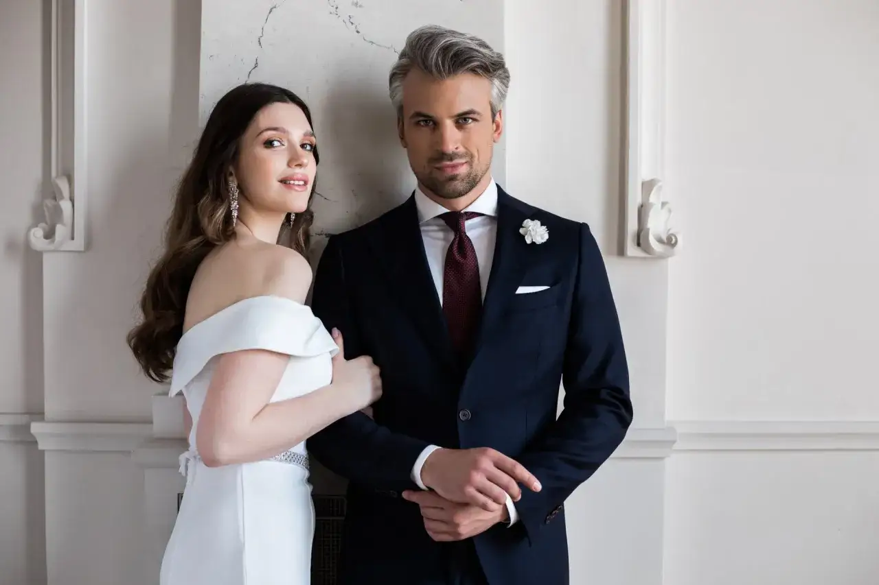 Elegancki mężczyzna w granatowym garniturze, białej koszuli i bordowym krawacie, idealny wybór jaki krawat do granatowego garnituru i białej koszuli na wesele. Obok niego kobieta w białej sukni.