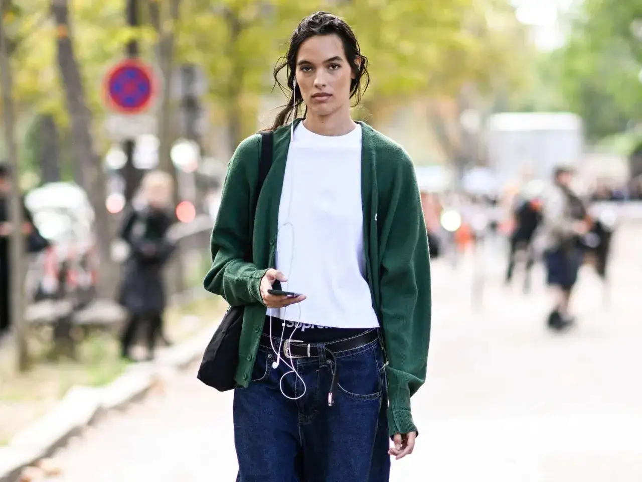 Kobieta w zielonym kardiganie i białej koszulce, z dżinsami i słuchawkami, prezentuje styl normcore.