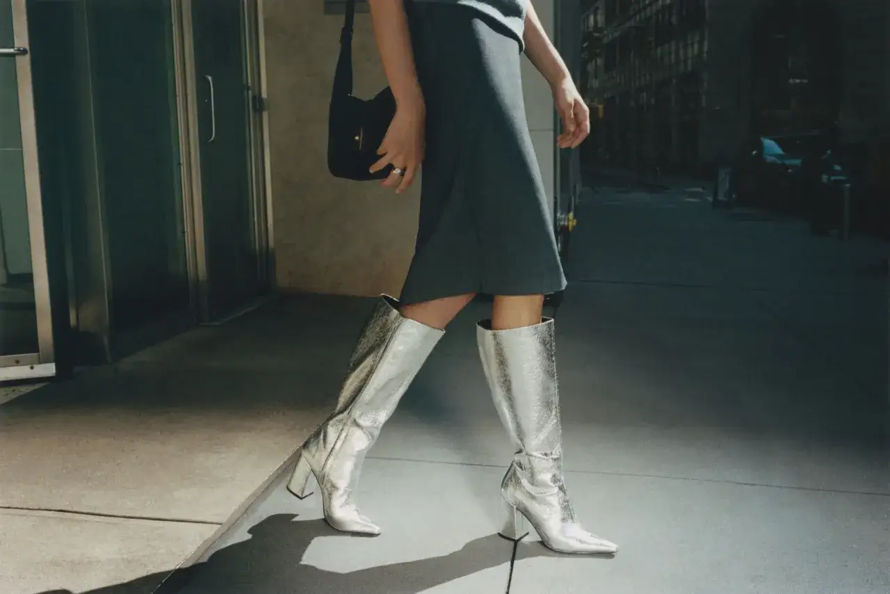 Kobieta w srebrnych kozakach i szarej sukience, z czarną torebką, prezentuje styl glam rock na tle miejskiego krajobrazu.