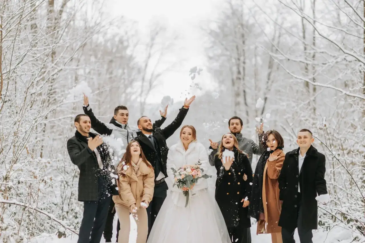 Radosna gromada rzuca śnieżkami w zimowym lesie. Panna młoda w białym futerku i pan młody w eleganckim płaszczu, otoczeni przez przyjaci&oacute;ł, tworzą piękne stylizacje na wesele zimą.