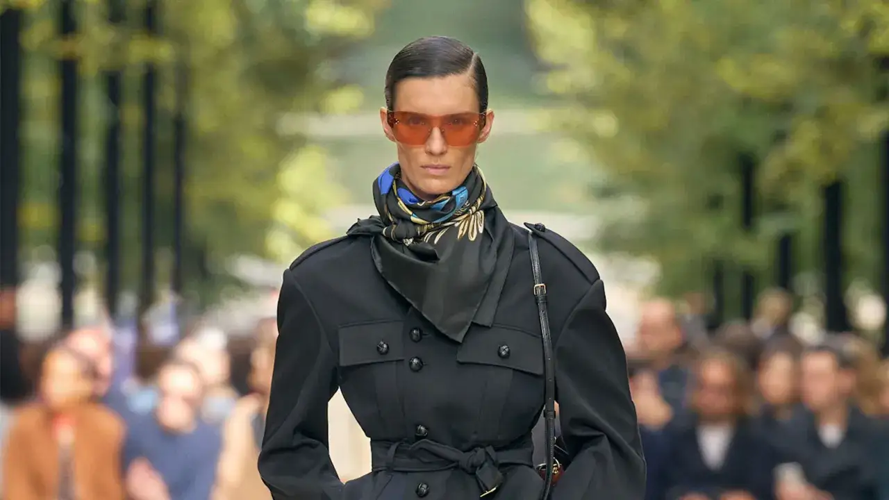 Modelka prezentuje eleganckie stylizacje wiosna 2026: czarny płaszcz z paskiem, apaszka i okulary w odcieniu pomarańczy.