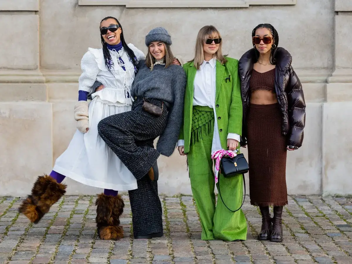 Cztery kobiety w ekstrawaganckich stylizacjach, prawdziwe fashion victim. Jedna w białej sukni i futrzanych butach, druga w szarym swetrze i spodniach, trzecia w zielonym garniturze, czwarta w brązowym komplecie.