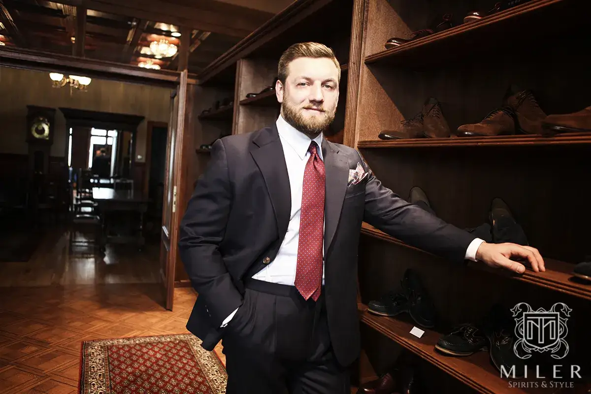 Elegancki mężczyzna w granatowym garniturze i czerwonym krawacie stoi w stylowym wnętrzu, obok półek z butami.