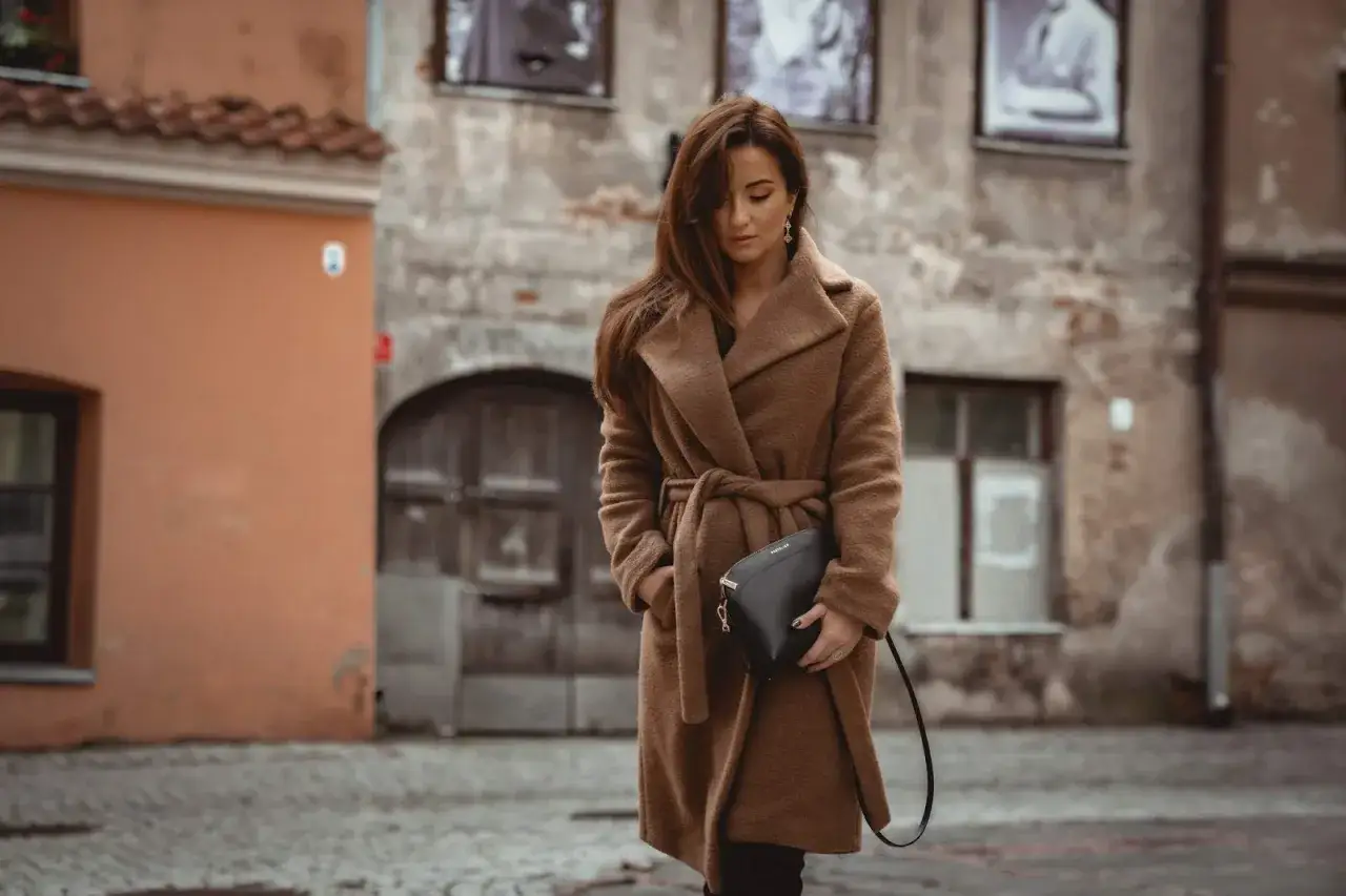 Elegancka kobieta w brązowym płaszczu z paskiem, trzymająca czarną torebkę. Idealne dodatki do brązowego płaszcza na jesienny spacer.