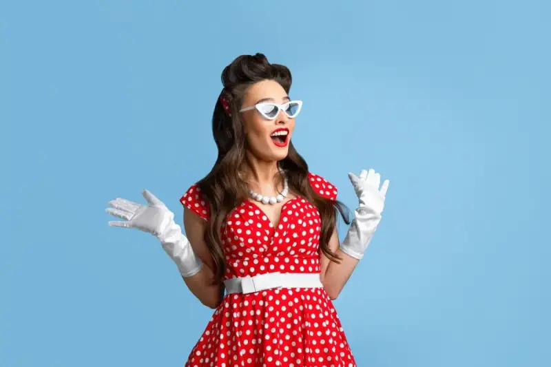 Urocza pin up girl w czerwonej sukience w grochy, białych rękawiczkach i okularach w kształcie kocich oczu, z uniesionymi rękami w geście zdziwienia.