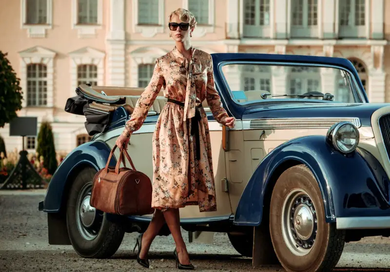 Elegancka kobieta w kwiecistej sukience i okularach przeciwsłonecznych, z brązową torbą podróżną, przy zabytkowym samochodzie. Styl vintage moda damska.